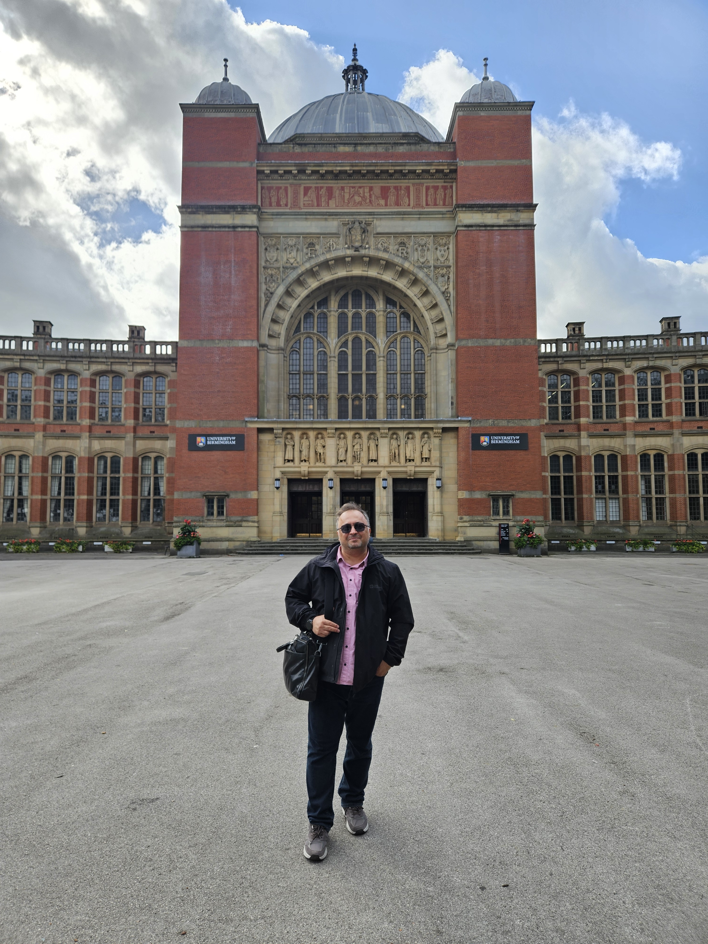 A man standing in a large paved courtyard in front of a red-brick university building with a domed clock tower and an arched entrance.