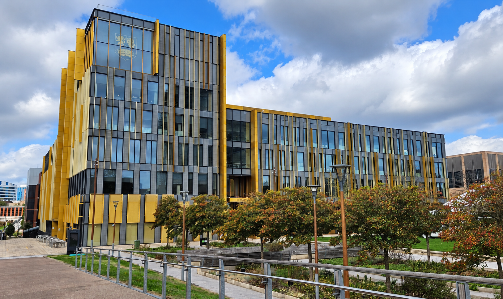 Modern university building with a glass-and-gold facade and an emblem above the entrance, under a partly cloudy sky.