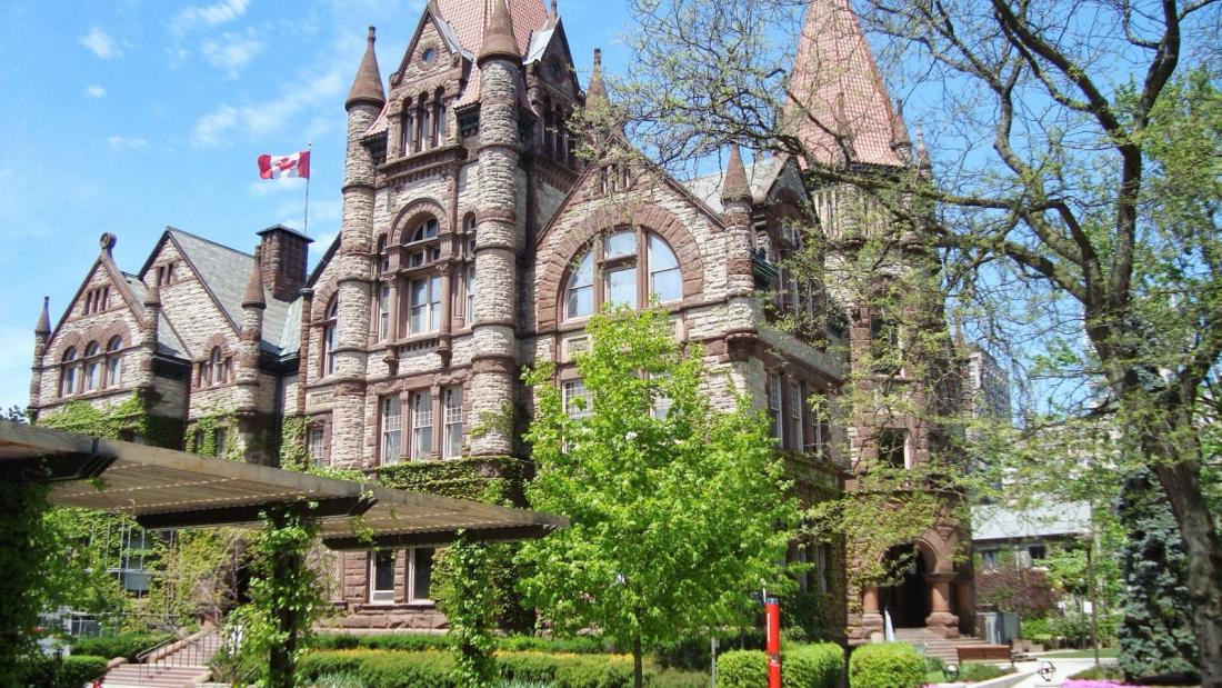 Historic stone building with small turrets and a Canadian flag, framed by trees.